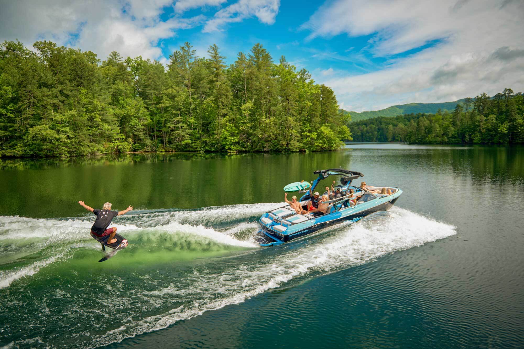 Wakeboarder riding behind a blue Malibu wakeboat on a forested lake