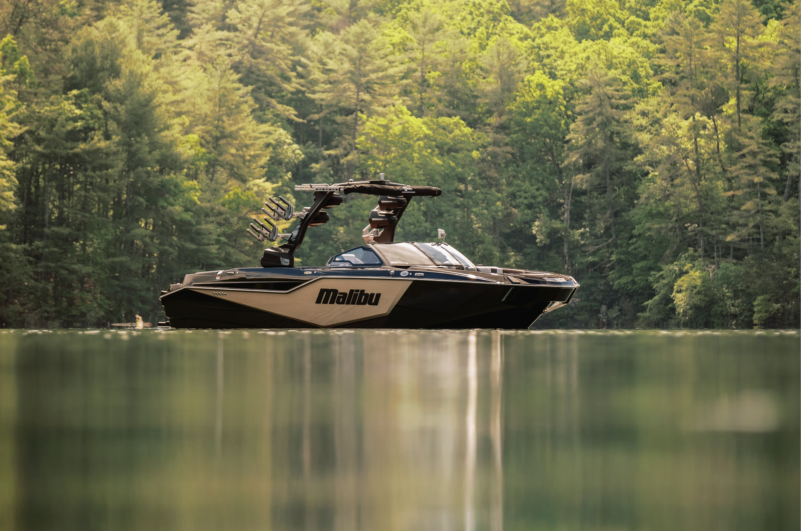 Black and gold Malibu wakeboat idling on a calm forest-lined lake