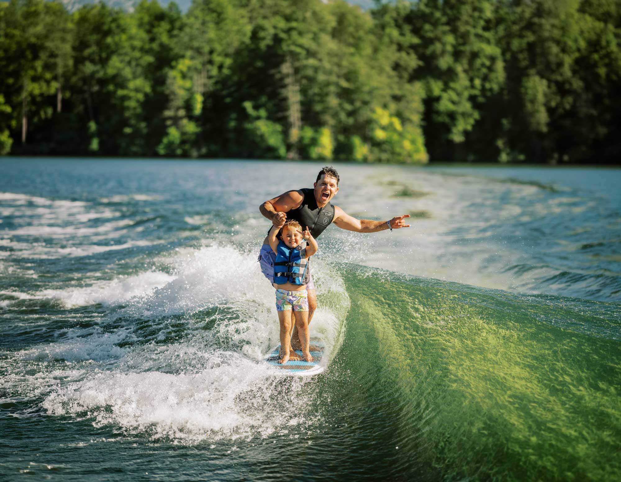 Father and child wakesurfing together on a wave behind a boat