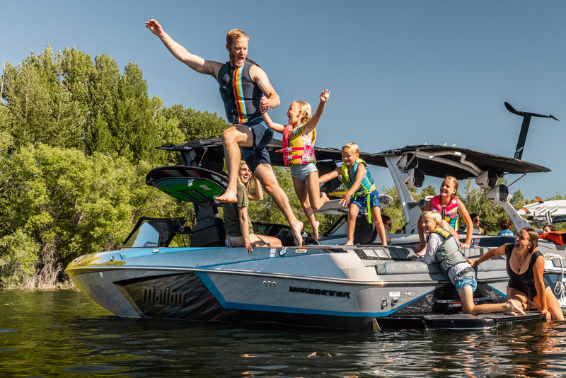 Family on a Malibu wakeboard boat on a lake, one person leaping off the bow into the water