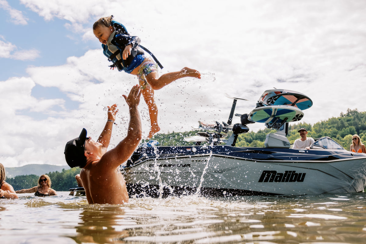 Father in the lake tossing his young child in a life jacket into the air beside a Malibu boat
