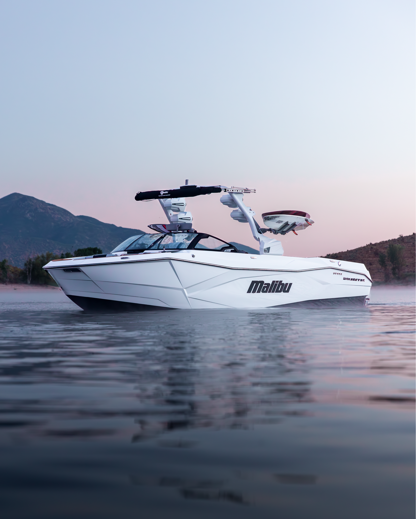 White Malibu wakeboard boat on a mirror-calm lake at dusk with mountains behind