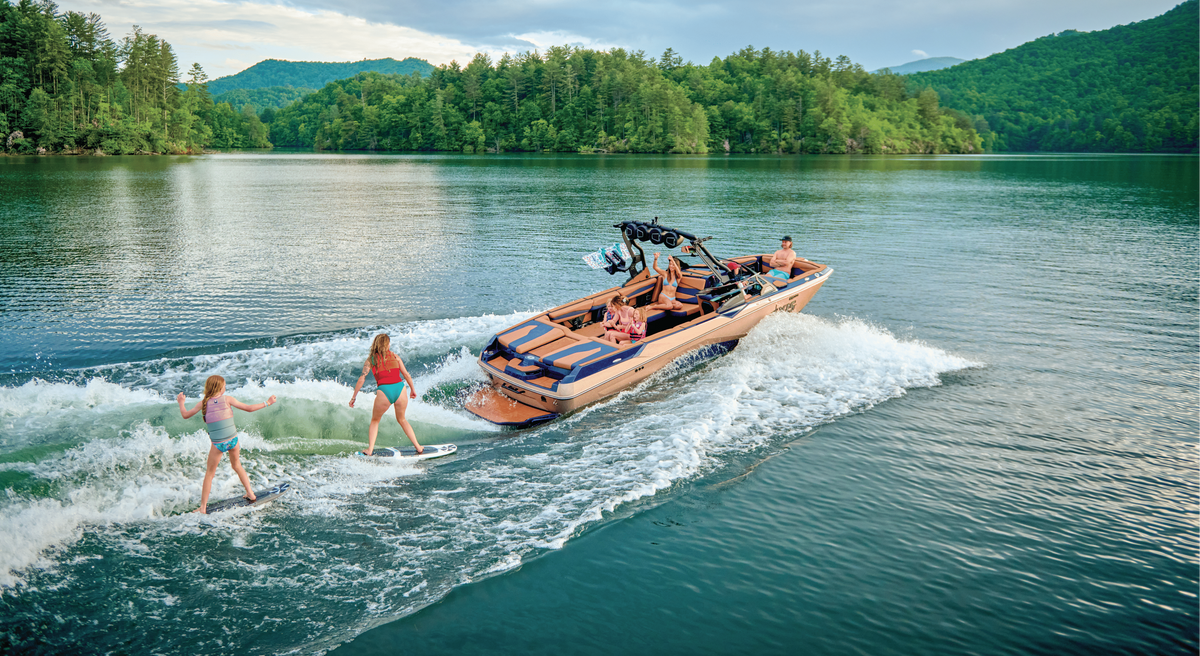 Black Malibu wakeboat towing two surfers across a forested lake