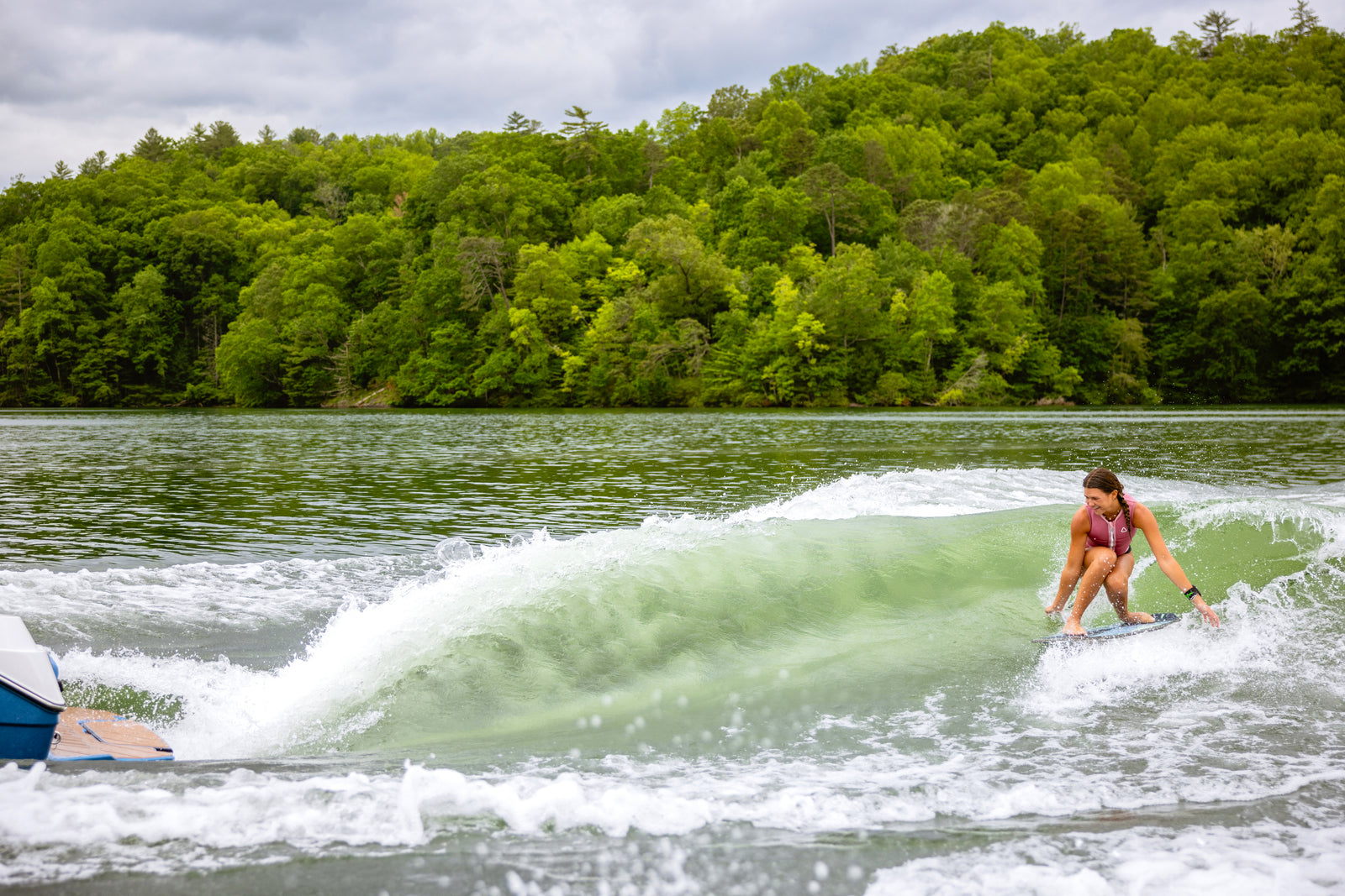 Woman in red swimsuit wakesurfing a clean wave behind a boat on a forested lake