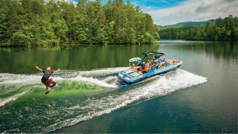 Blue Malibu boat towing a wakeboarder grabbing air on a tree-lined lake