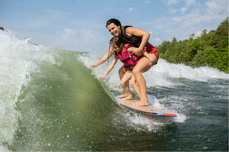 Two women wakesurfing the same wave together behind a boat