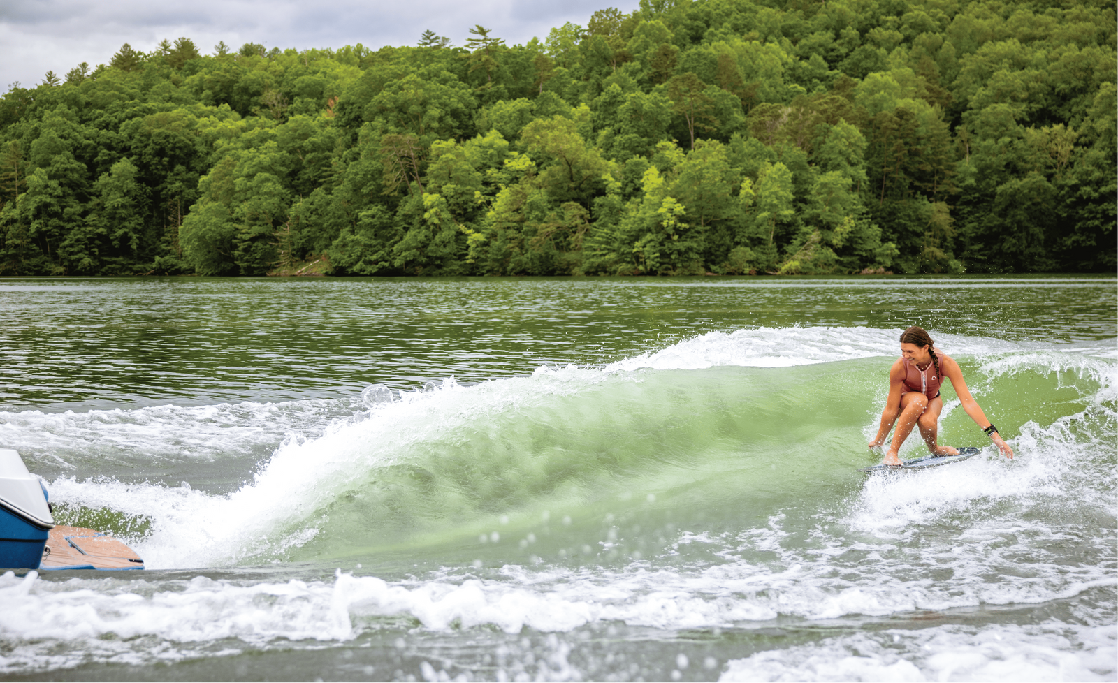 Wakesurfer carving a green wave behind a boat on a forested lake